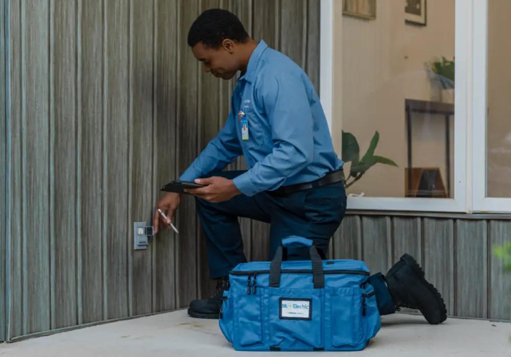 Technician in blue uniform kneeling beside a gray exterior wall, inspecting an outdoor electrical outlet with a pen and tablet; blue tool bag on ground.